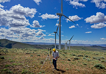 A surveyor with tripod and telescope, 戴着黄色头盔, taking notes with wind turbines in background